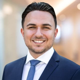 Professional headshot of a smiling man in a dark blue suit and blue tie against a blurred office background
