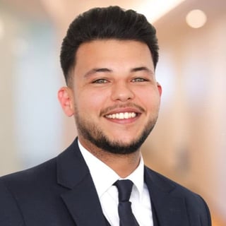 Professional headshot of a smiling man with dark hair and beard wearing a black suit and tie against a blurred background